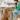 A woman in a green cardigan and blue jeans sits on a chair at a kitchen island, holding the paw of a fluffy golden-brown dog. Several colourful supplement jars from North Hound Life are displayed on the marble countertop behind them.