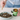 A person's hands are shown sprinkling a white powder from a small wooden spoon into a pet puzzle bowl filled with kibble. Next to the bowl is an amber glass jar of North Found Life Whole Health dog food supplement with a green label.